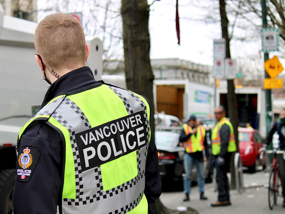 A man stands on the sidewalk in the Downtown Eastside wearing a yellow vest that says 'Vancouver Police.' Other people wearing safety vests, and one person riding a bike, are visible in the background. 