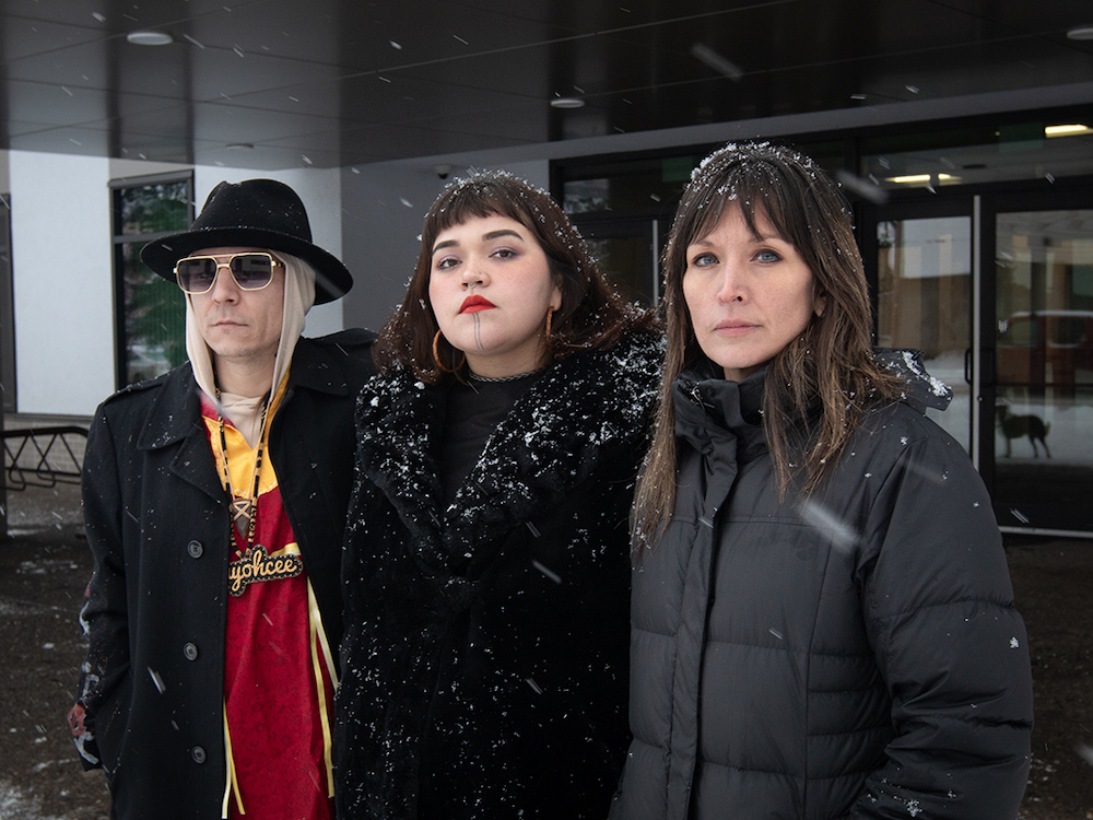 Three people stand close together outside the entrance of a building. The man on the left is wearing sunglasses and a hat. Two women with dark hair wear all black. Snow is falling.