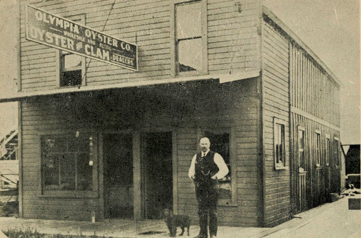 An old black and white photo shows a man in a waistcoat, accompanied by a dog, standing outside an oyster and clam dealer building.