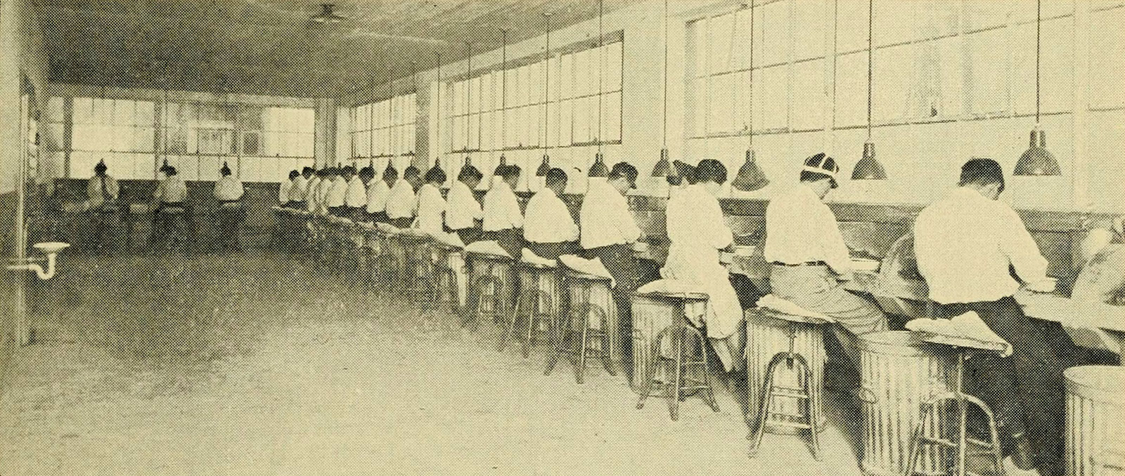A black and white photo shows a couple of dozen people sitting on stools in front of high counters, shucking oysters.