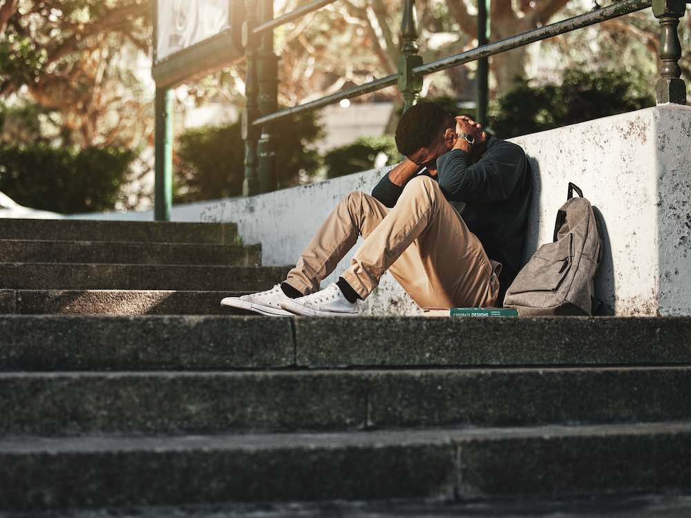 A young Black man sits on outdoor stairs, his hands beside his head in a gesture of frustration and despair. A day pack is beside him, and he wears white sneakers, brown pants and a dark sweater.