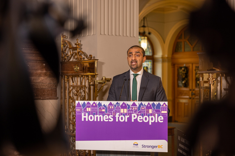 A man with a green tie makes an announcement in an ornate legislative building in front of a podium with a purple poster that says 'Homes for People.'