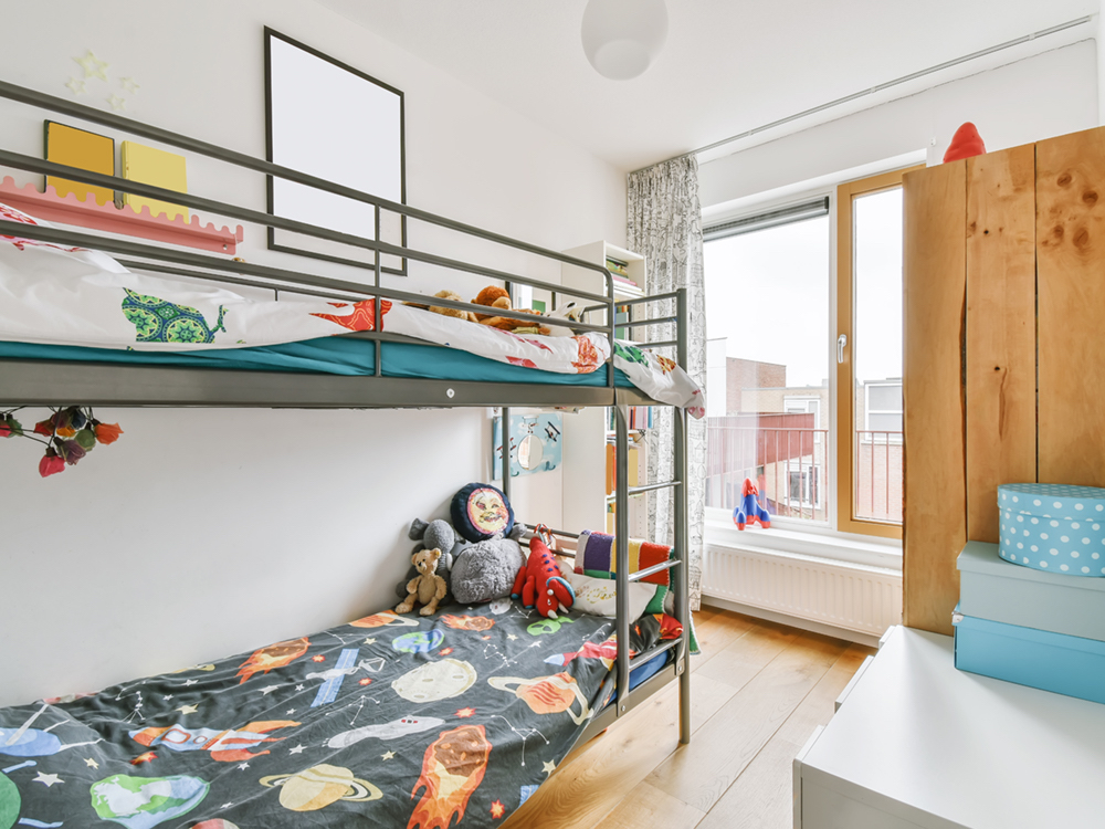 A metal bunk bed stands in a small, white-walled room with hardwood floors. At the end of the room is a large window and a glass door to a patio.