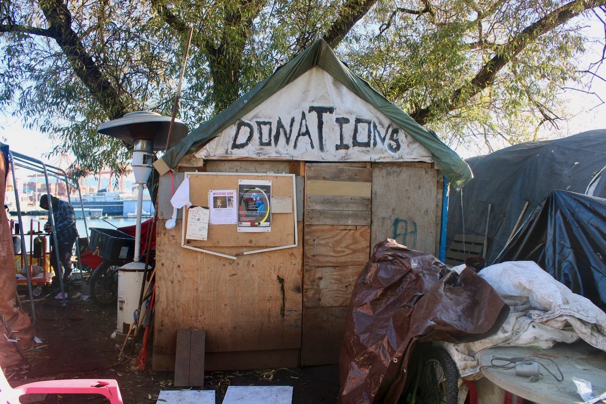 A plywood shack with a green tarpaulin roof stands under a tree. The word 'Donations' is scrawled over the door in large black letters.