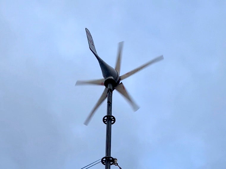A wind turbine with a long slender grey body and five lighter-coloured blades spins in a blue sky.