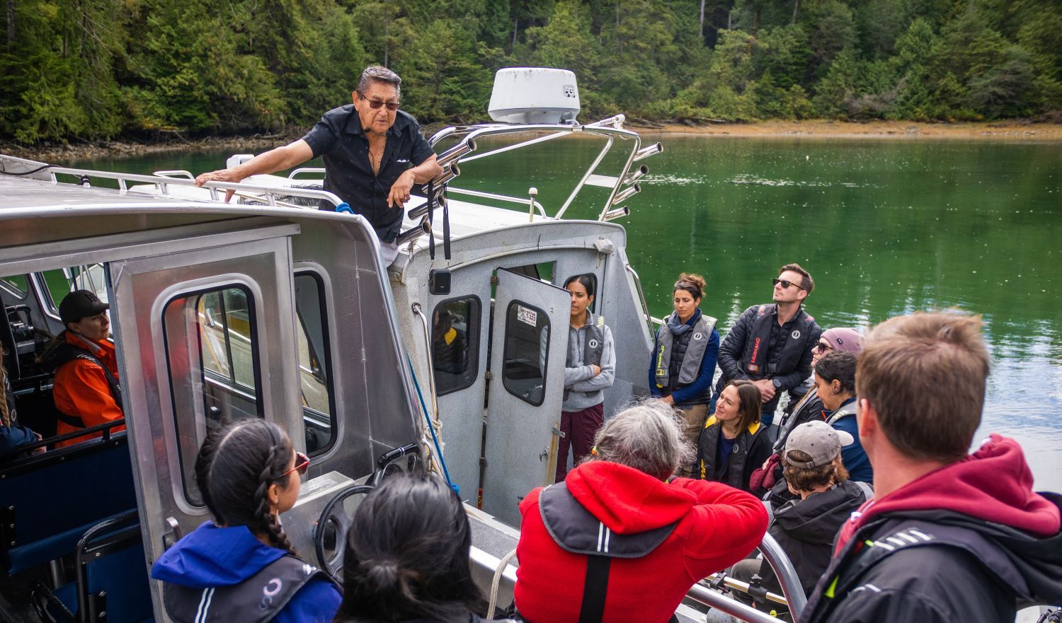 An older man stands between two boats, above a number of students who are in the boats and paying attention to what he's saying.