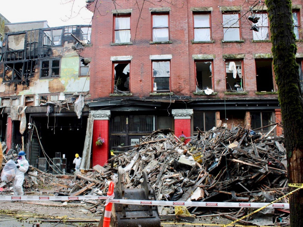 A fire scene, with piles of rubble, broken windows in a red brick building and almost destroyed upper floor. A digger and people with white biohazard suits are on the scene.