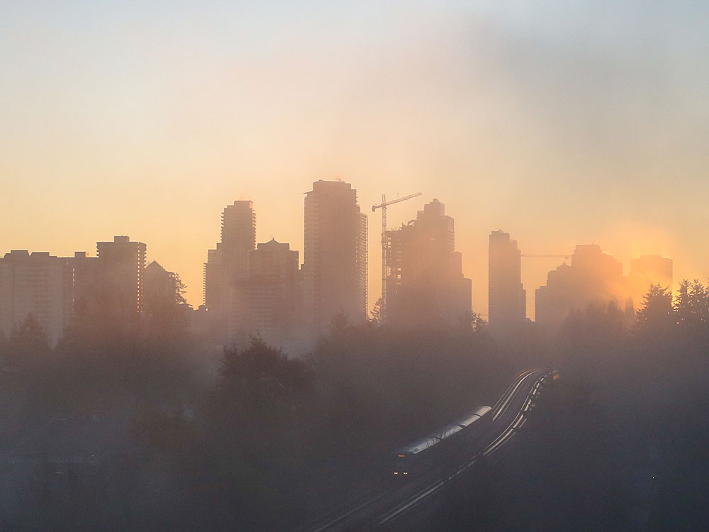 The sun rises behind a skyline in the fog. From between the towers a track emerges with a train on it.