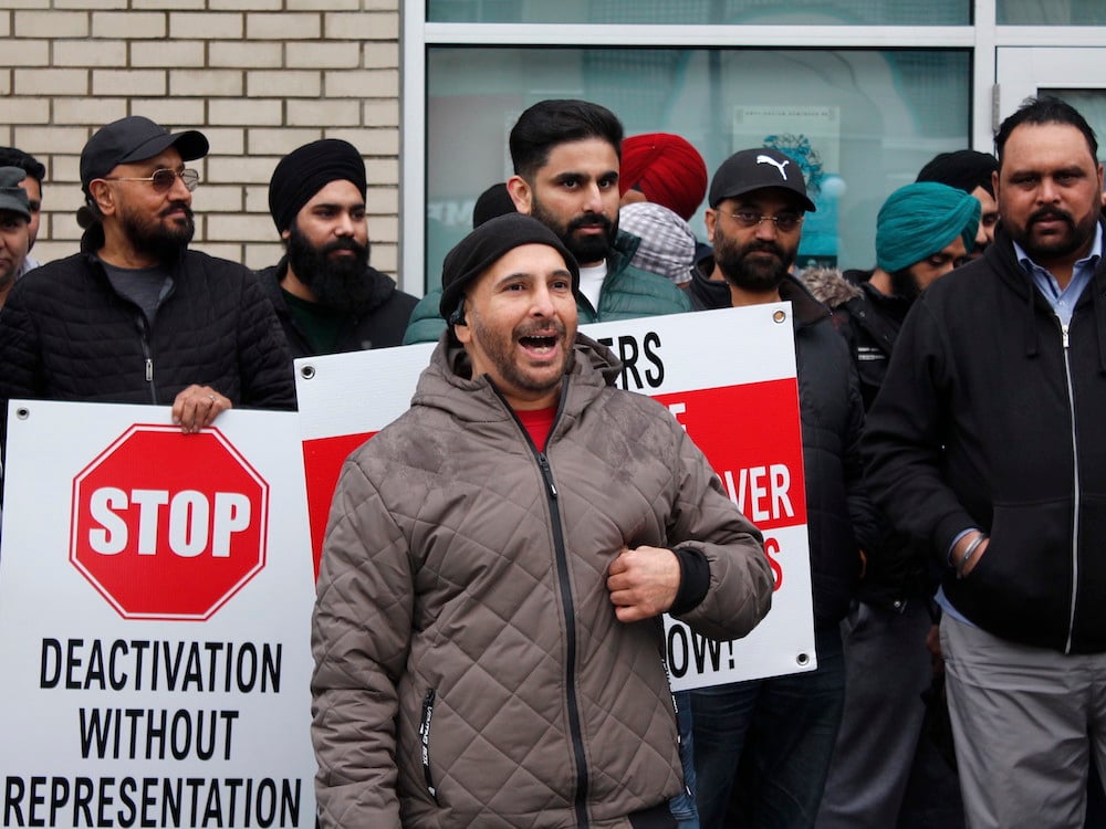 An Indo-Canadian man in a brown jacket and black toque is smiling and speaking in front of a group of other Indo-Canadian men. A sign to his right says 'STOP DEACTIVATION WITHOUT REPRESENTATION.'