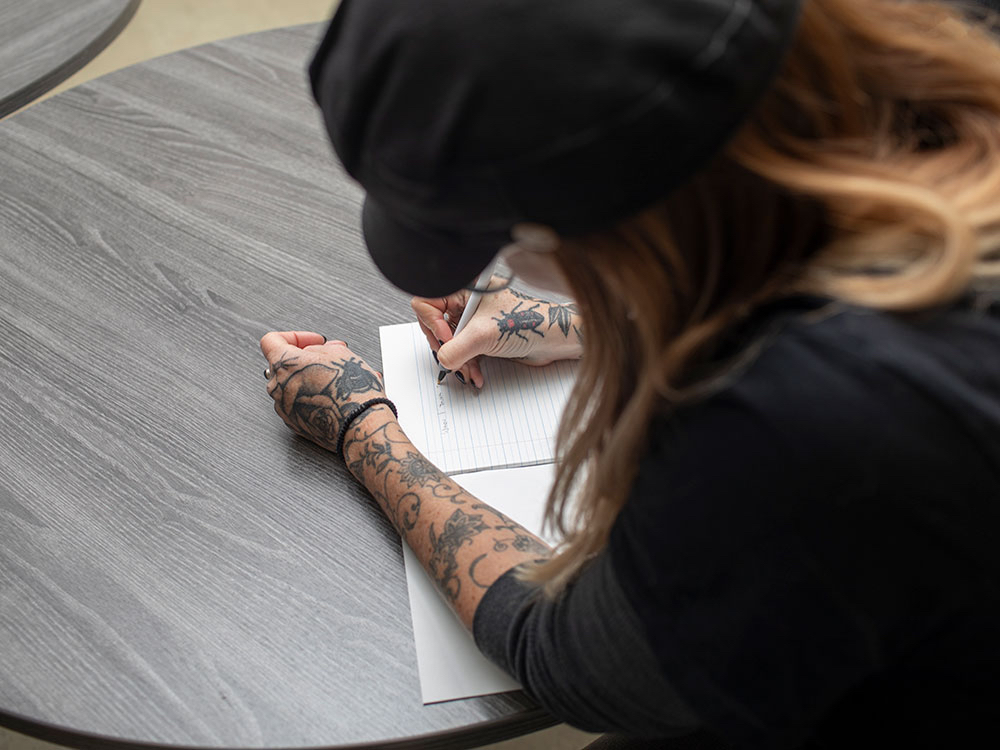 A person in a black cap with tattooed arms sits at a grey round table holding a pen and writing in a looseleaf notebook.