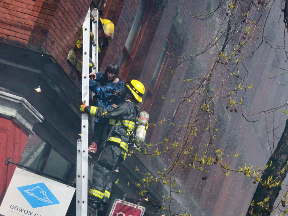 A firefighter in safety gear stands on a ladder, while above another helps a woman out a hotel window to safety as smoke hangs in the air. She is wearing a blue plaid shirt or blanket and appears to be gasping for air.