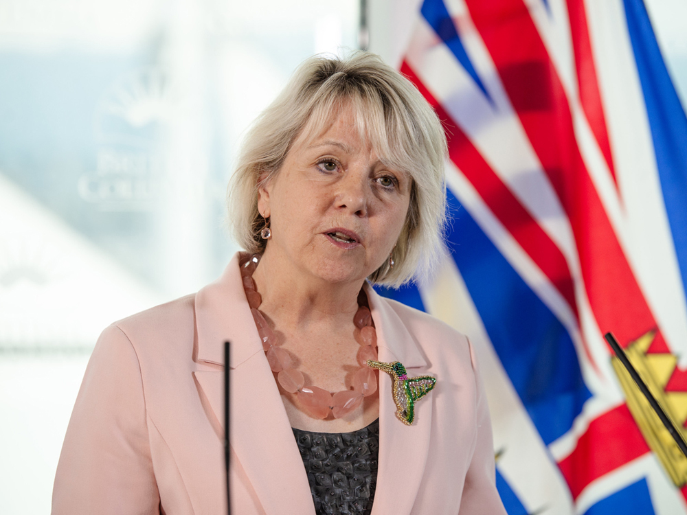 Bonnie Henry, a middle-aged white woman with light blonde shoulder-length hair, stands at a podium with the B.C. flag in the background. She is wearing a pink blazer and a beaded hummingbird pin.