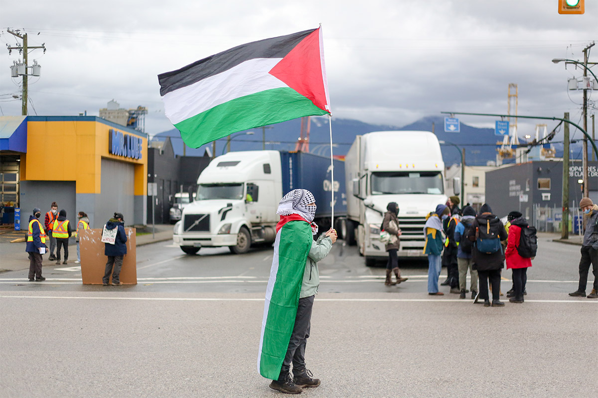 A person stands on a road while flying a Palestinian flag and wearing a second flag over their shoulders. They are also wearing a Palestinian scarf over their head and face. There are transport trucks and people milling around in the background.