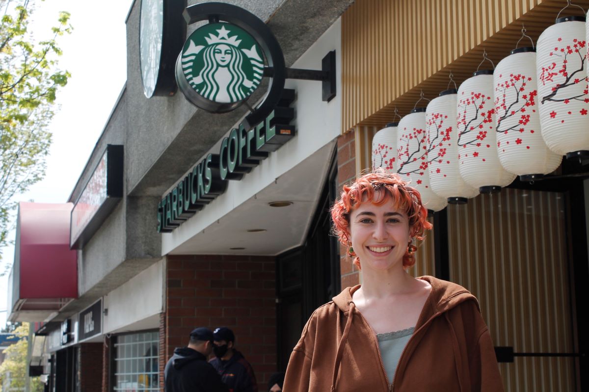 A woman with light skin and short wavy red hair, wearing a brown hoodie over a light green shirt, stands outside a Starbucks.