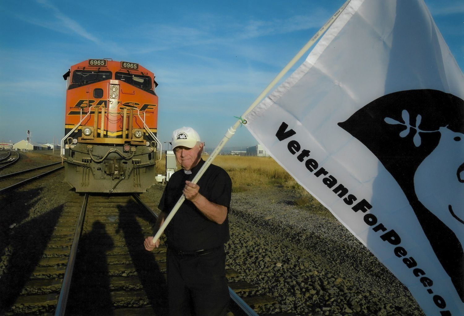 A white man wearing a white ball cap and a short-sleeved black shirt with a reverend's collar stands on a railroad track with an orange train close behind him. He is holding a white flag that says 'VeteransForPeace.org.'