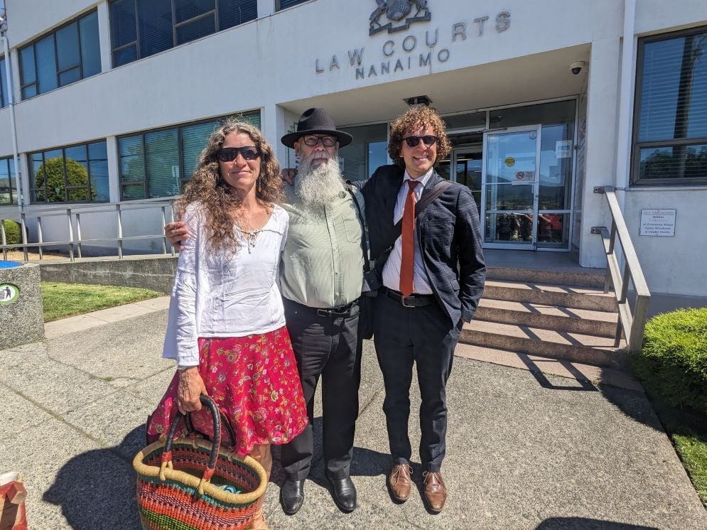 Three people stand in front of the Nanaimo Law Courts building. From left, a woman with light skin and long curly brown hair wearing a white peasant blouse and red patterned skirt; a man with light skin and a long grey beard wearing a black fedora, a moss green button-up shirt and black pants; and a man with light skin and curly brown hair wearing a dark casual suit and red tie.