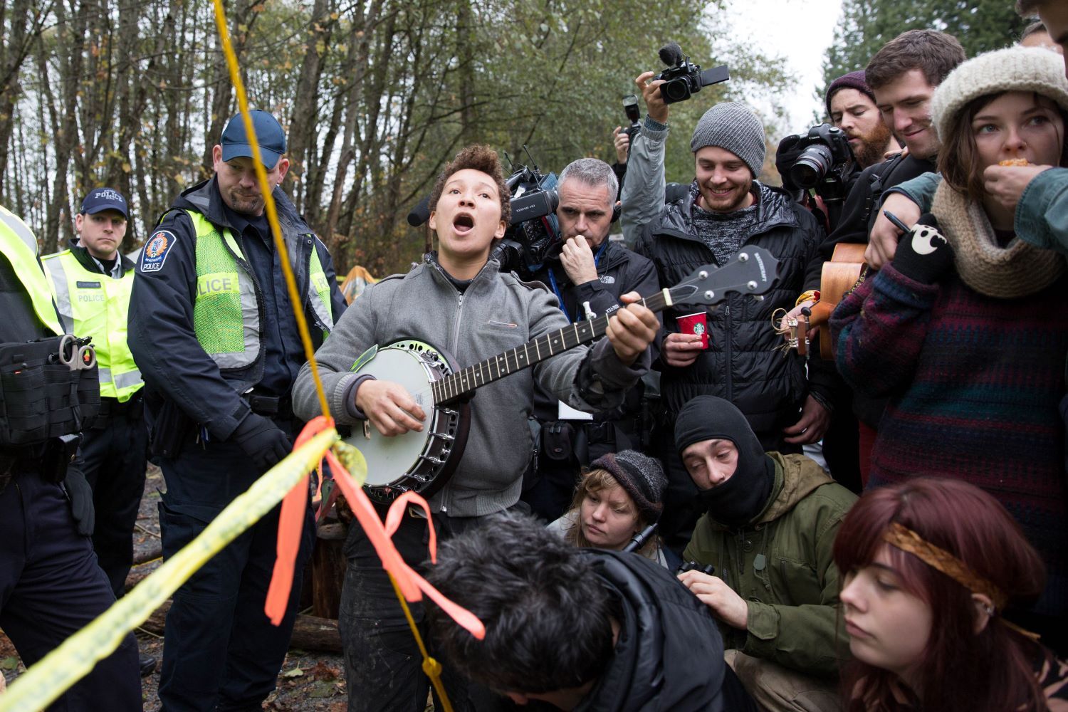 A mixed group of police officers, young people and photographers at a protest site. In the centre is a young person playing a banjo and singing.