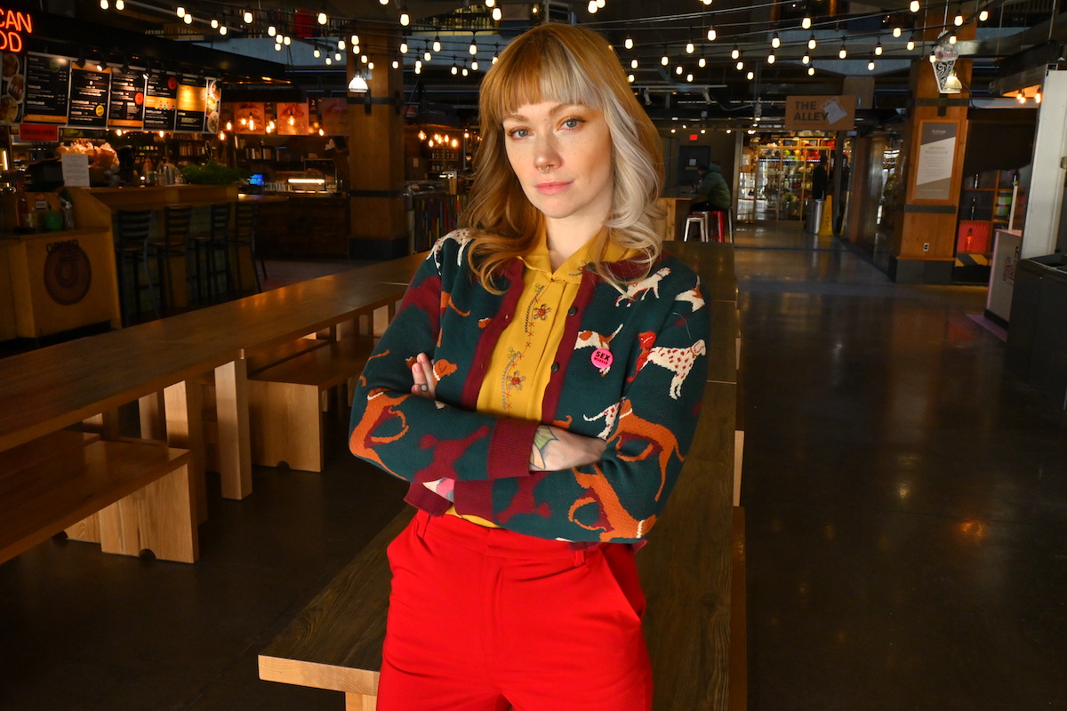 A white woman with orange and blond hair poses with her arms crossed. She is wearing red pants and a cardigan festooned with dogs.