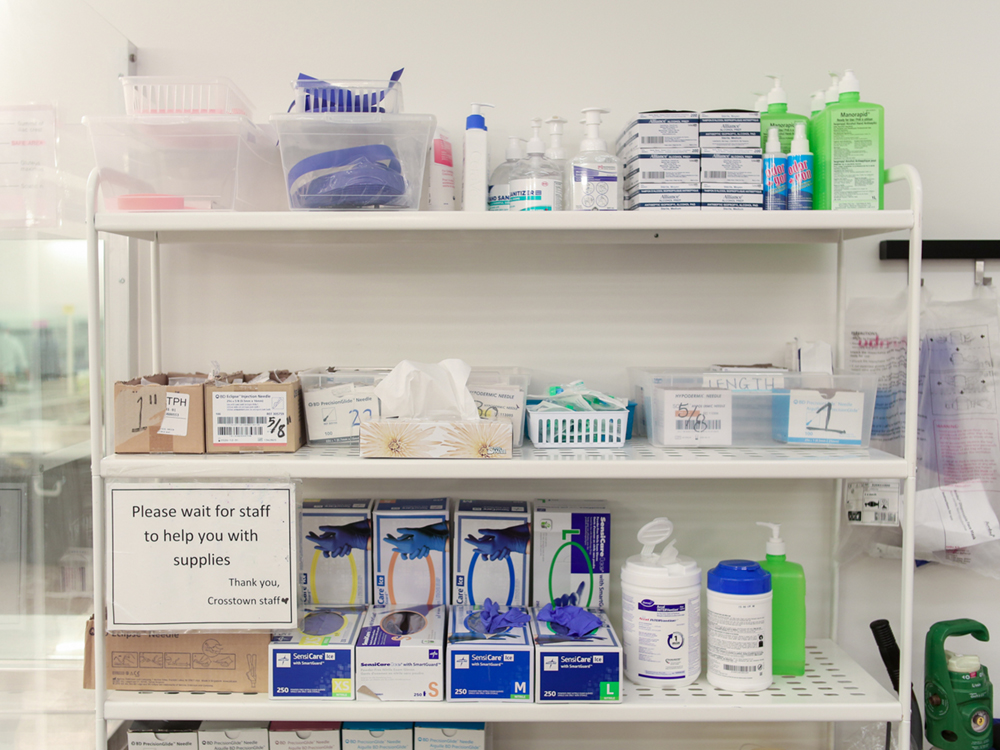 Harm reduction supplies on a shelving unit. They are well-organized and labelled. A sign says ‘Please wait for staff to help you with supplies.’