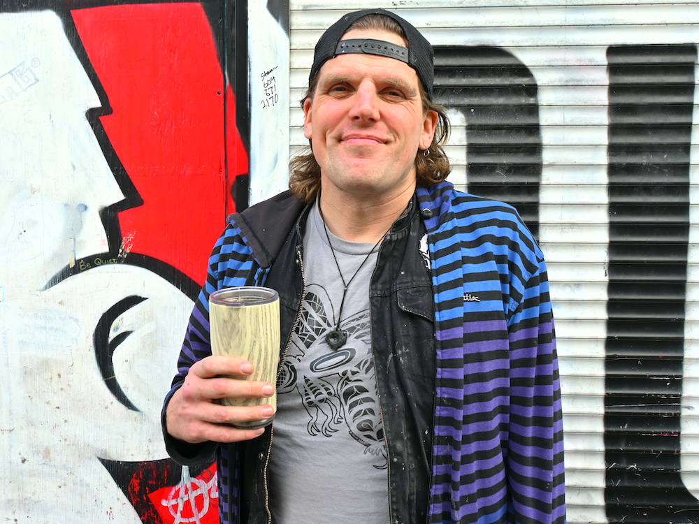 A man with light skin and shoulder-length wavy brown hair wears a backwards cap, black jean jacket and hoodie. He holds a travel mug of tea.