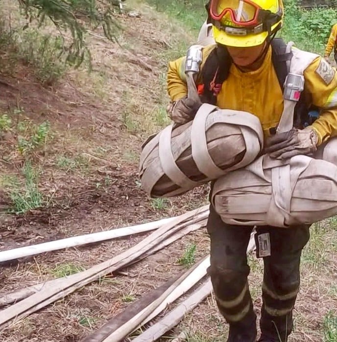 A person in yellow safety gear and helmet carries two large bundled houses slung over their shoulders.