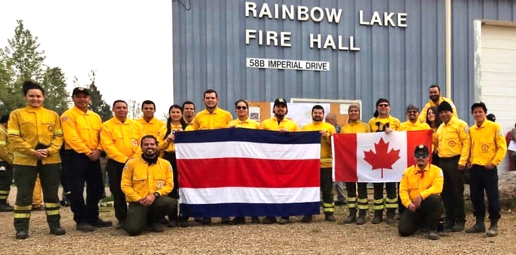 A large group of people wearing matching yellow shirts stand outside a metal building with a sign that says 'Rainbow Lake Fire Hall.' Several of them are holding up a Costa Rican flag, others a Canadian flag.