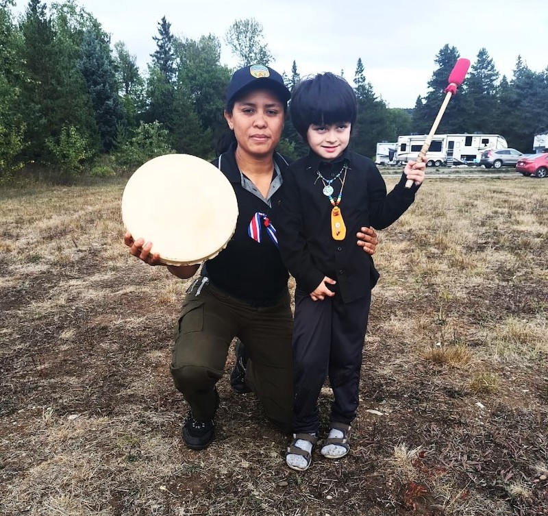A woman with medium skin tone and black hair holds a drum and poses with a young, dark-haired boy wearing decorative necklaces, holding a drum-beating mallet and smiling shyly.