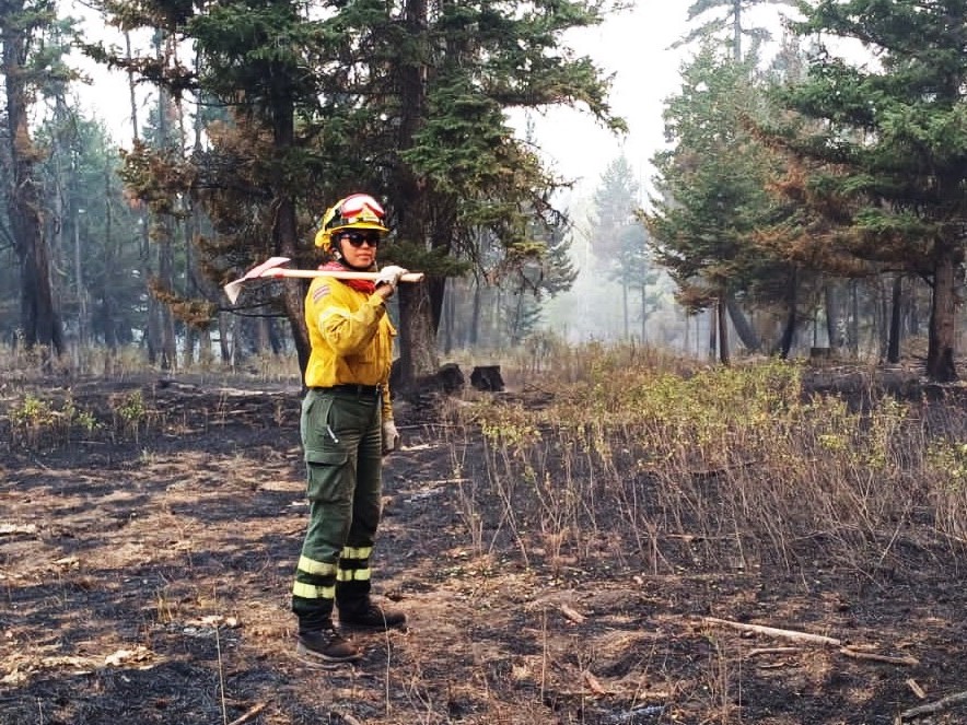 A woman stands on a patch of burned ground, wearing boots and safety gear including a yellow jacket and safety helmet. She looks toward the camera and carries a large axe on her shoulder. Smoke rises in the background.