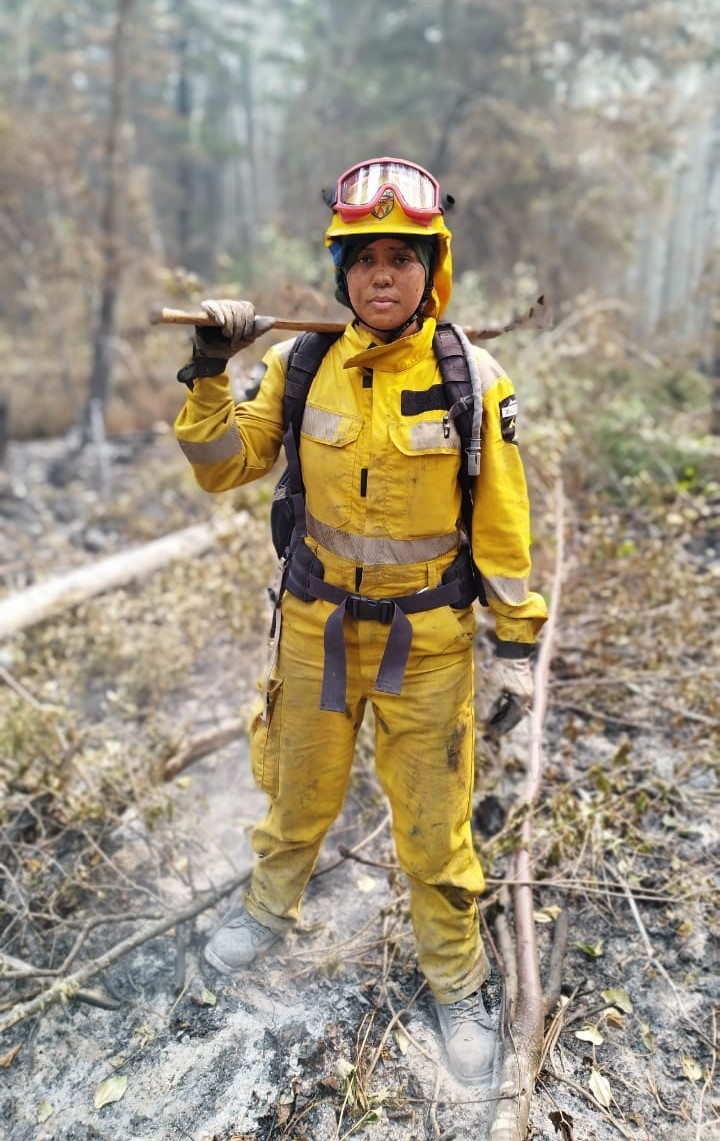 A Costa Rican woman wearing yellow safety gear and goggles perched on her helmet stands with trees behind and white ash at her feet.
