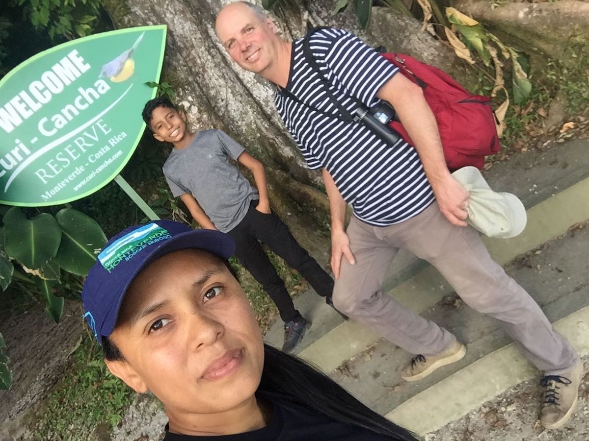A Costa Rican woman takes a selfie in front of a sign welcoming visitors to the Curi-Cancha nature reserve. Behind her a boy smiles, wearing a grey T-shirt and dark pants. Beside him, a light-skinned man smiles at the camera, wearing a black-and-white striped shirt and wearing a red backpack.