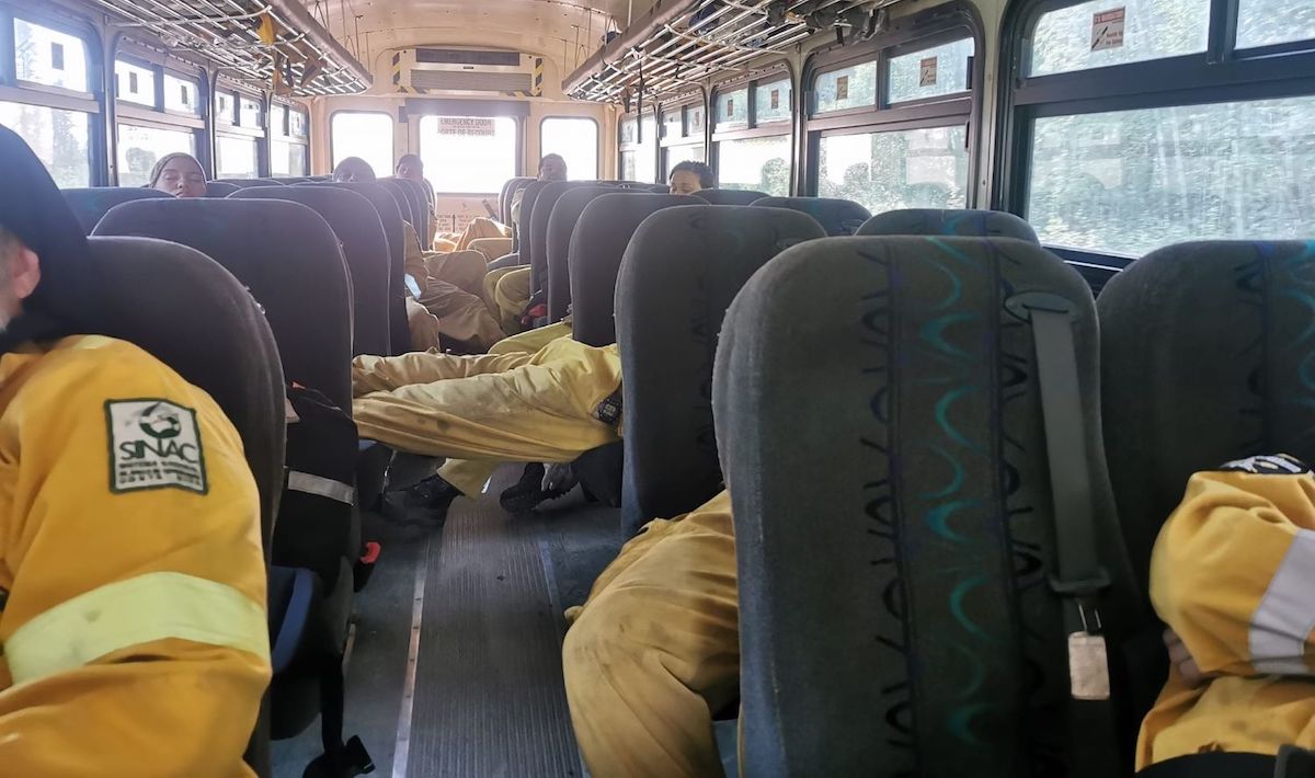 Exhausted workers in yellow safety gear are sprawled on the seats of a bus, some lying down.