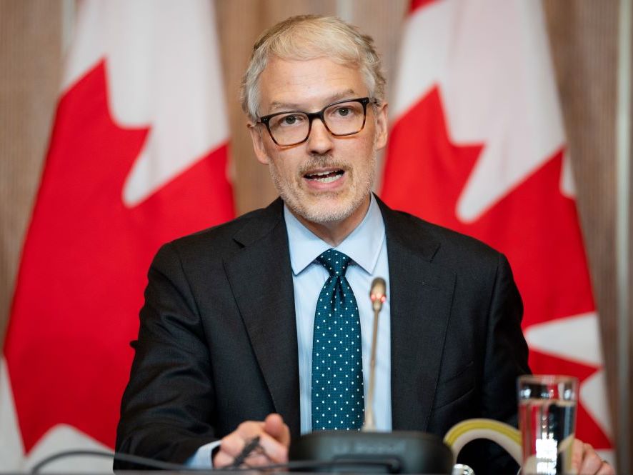 A white-haired white man with black-rimmed glasses speaks into a microphone with the Canadian flag behind him.