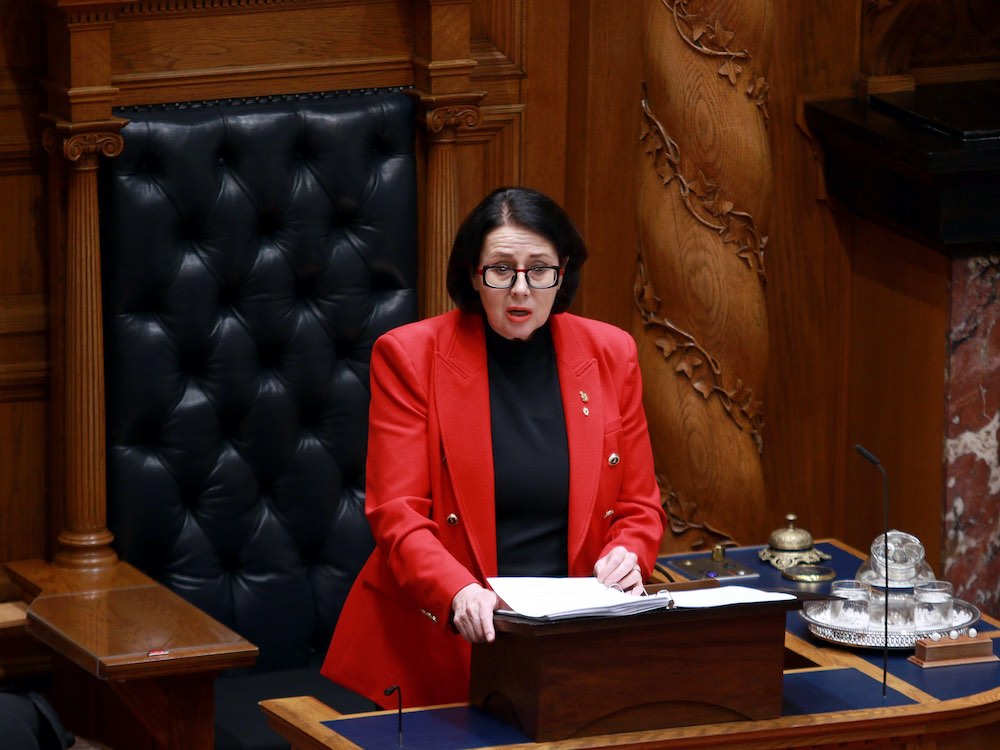 A middle-aged pale woman with dark hair, a red jacket and a black shirt stands at a podium reading a speech. Behind her is wood panelling and a large, black leather-covered chair.