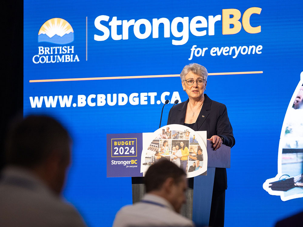 A grey haired, light-skinned woman stands at a podium speaking to media. Behind her white letters on a blue wall say ‘Stronger BC for everyone.’