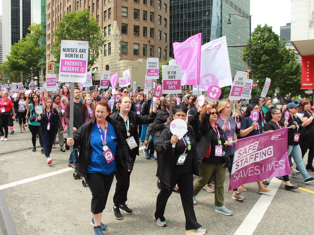 Nurses wearing pink lanyards and white, pink and black signs march down a Vancouver street.