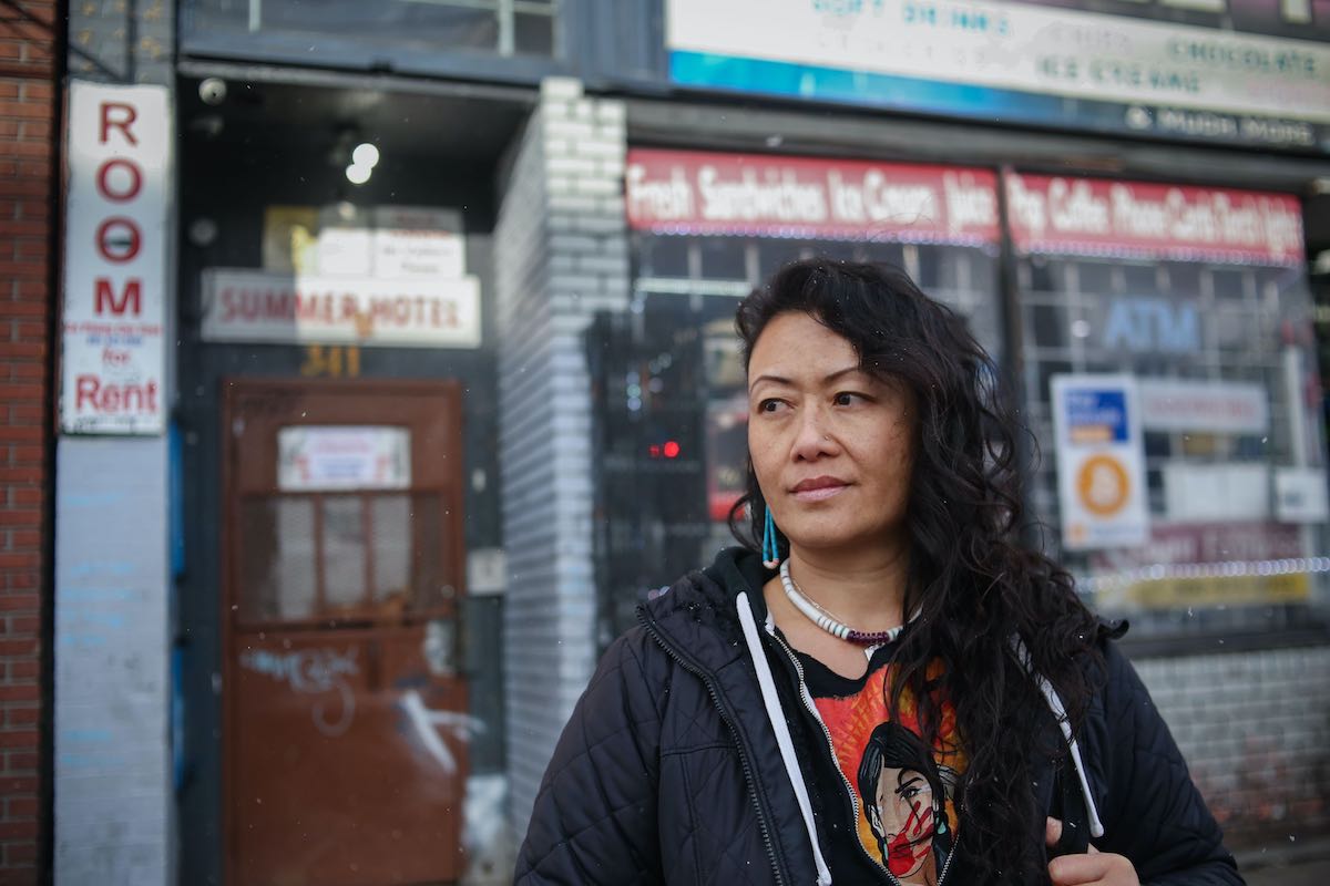 A medium-skinned woman with long black hair, wearing a hoodie and beads, stands outside a rundown-looking hotel.