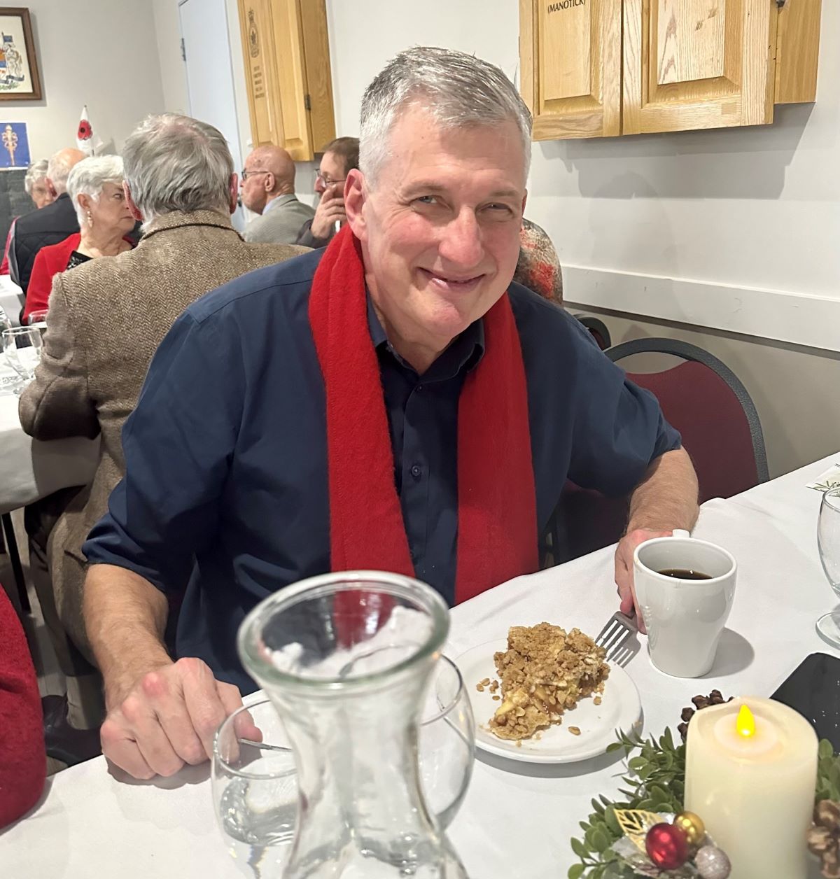 A light-skinned man with short grey hair wearing a navy shirt and red scarf sits at a table in a restaurant with a plate of apple crumble in front of him.