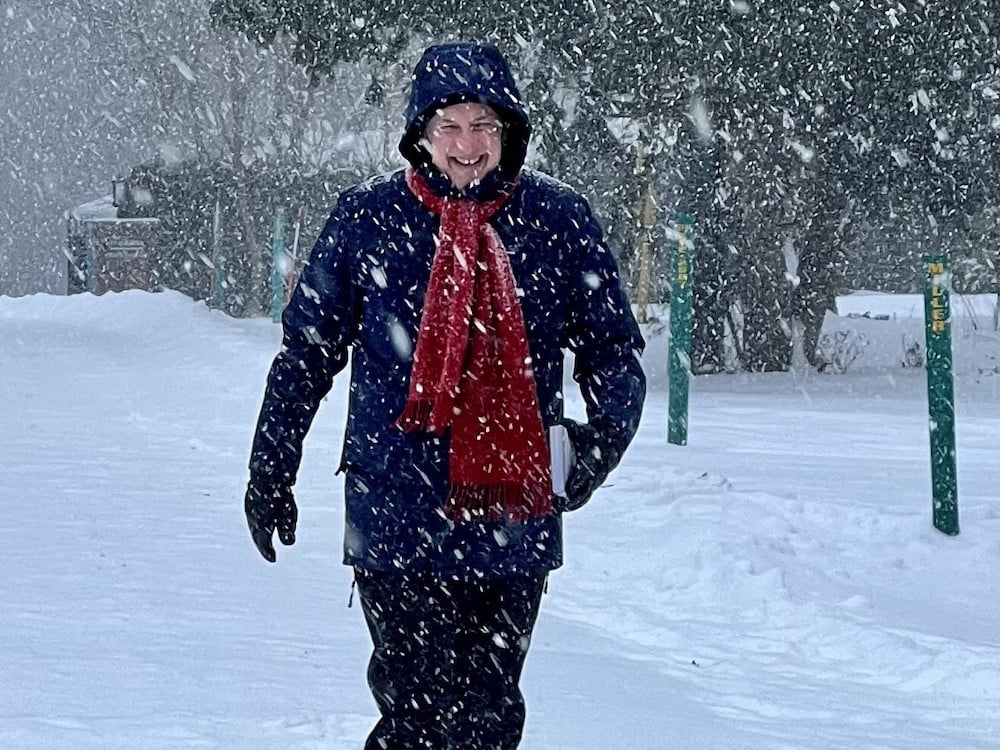 A light-skinned man in a blue winter coat and red scarf trudges through a blizzard with a big smile on his face.