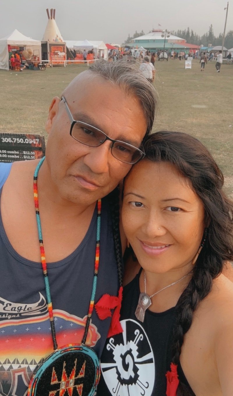Two people with medium skin tone pose with their heads together at an outdoor gathering. He wears beads and glasses; she has long, dark, braided hair.