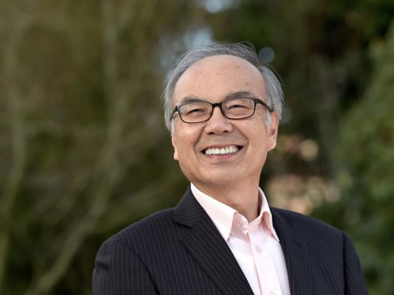 A man with short grey hair, glasses, a light shirt and pin-striped suit smiles at the camera.