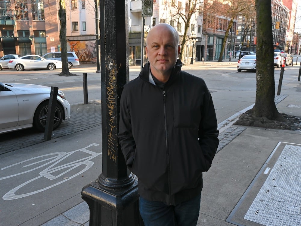 Thomas Kerr, a bald white man wearing a black jacket, leans against a light pole next to a street in Gastown, Vancouver.