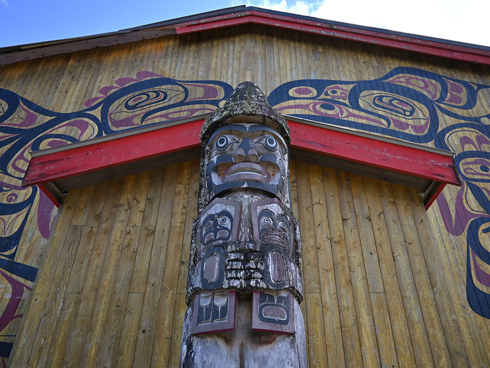 A carved and painted wooden totem pole in front of a painted wooden longhouse in Klemtu.