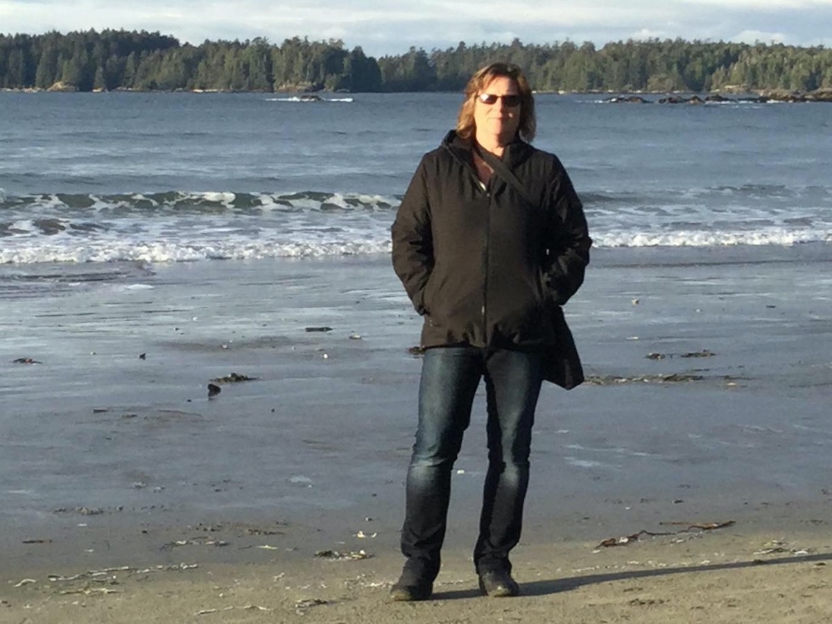 A woman in a black coat and blue jeans stands on a beach, hands in pockets. She has shoulder-length brown hair and the ocean is behind her.