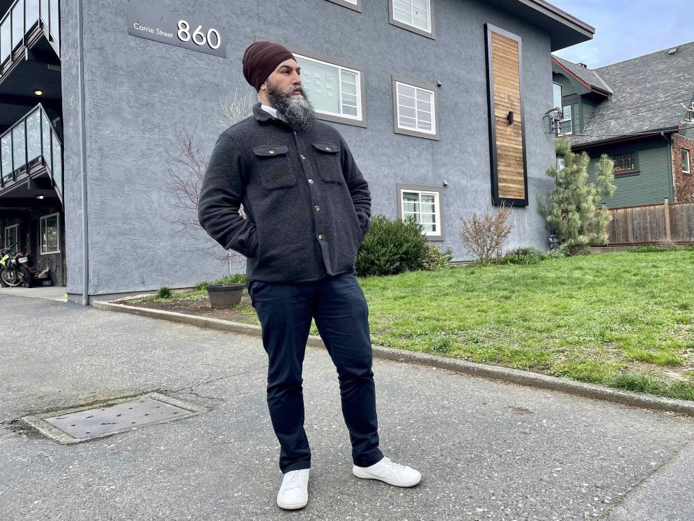 A man with medium skin tone wearing a burgundy turban, dark casual clothes and white shoes stands outside a grey apartment building with his hands in his pockets.