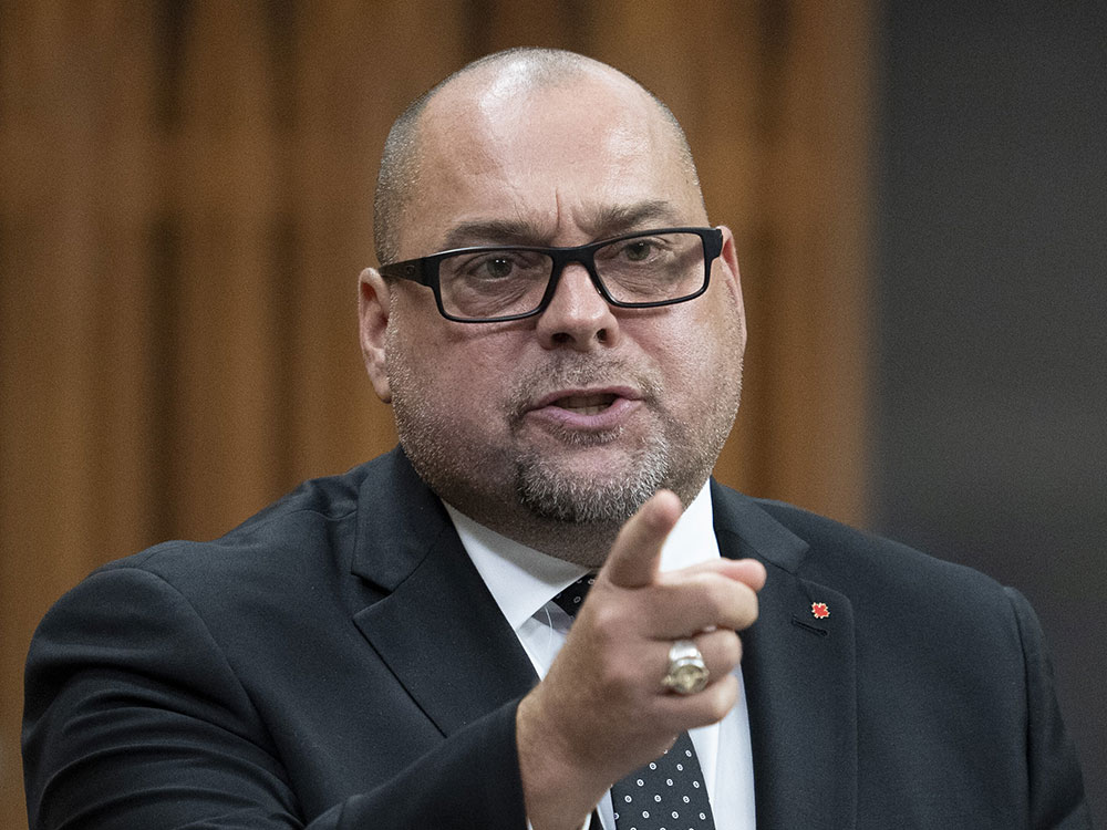 A bald white man wearing a dark suit, a close-cropped beard and dark rimmed glasses points with a fierce expression while standing in the House of Commons. 