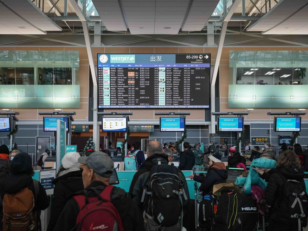 Passengers wait in the Vancouver airport, looking up at a large screen displaying flight schedules.
