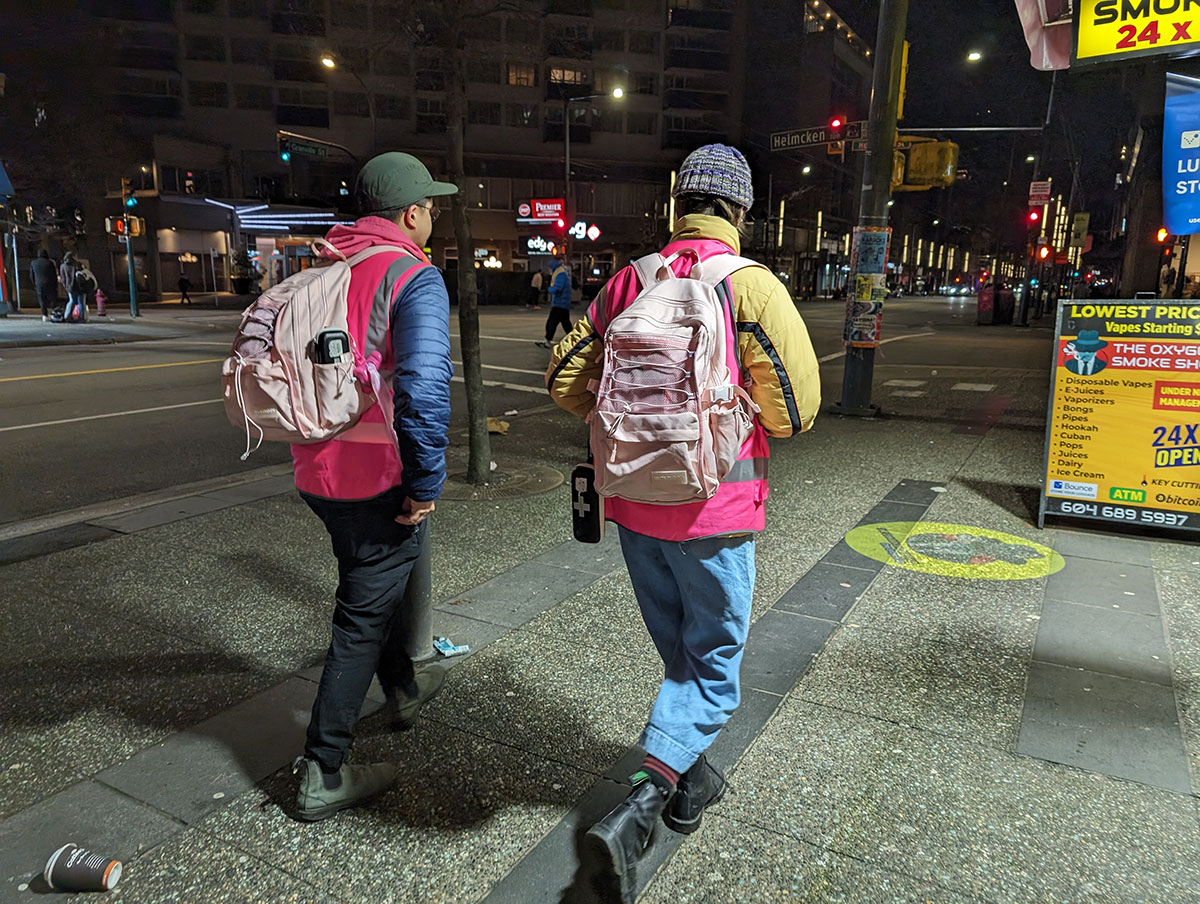 Two people in pink vests and pink backpacks walk down Granville, facing away from the camera.