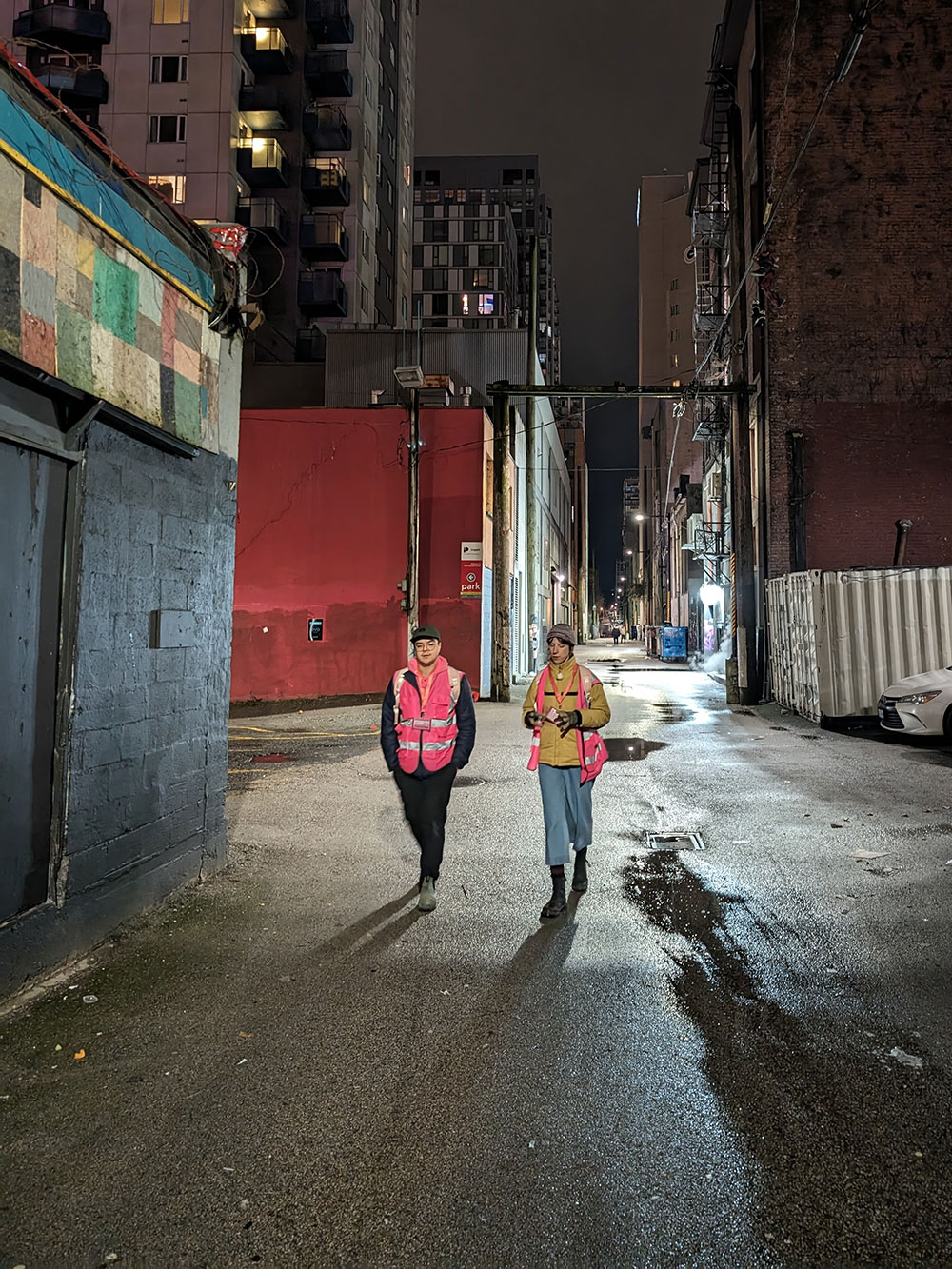Two people in pink vests walk down an alleyway towards the camera.