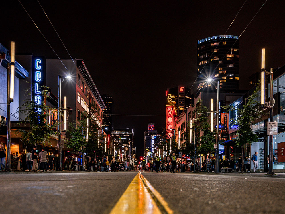 The Granville strip at night, taken from the middle of the road. The strip is full of brightly lit signs and is lined with partygoers.