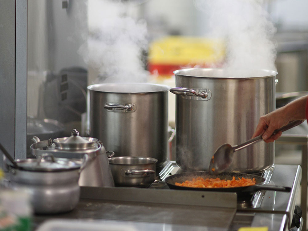 A stainless steel stove in a commercial kitchen shows two steaming hot pots, and a pan. A hand is stirring some kind of noodle dish.